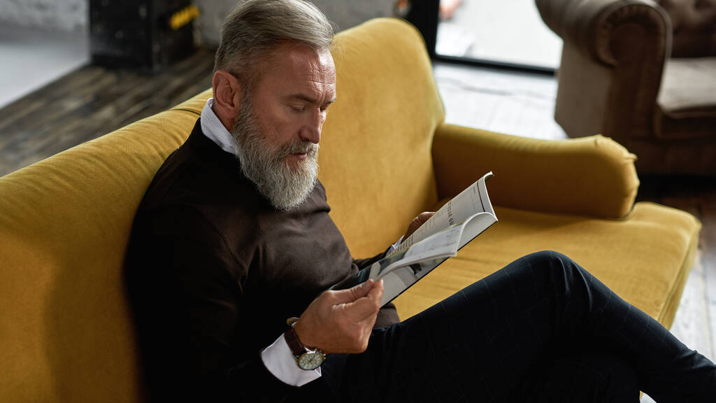 Senior concentrated caucasian man reading journal on sofa in spacious apartment. Stylish bearded pensioner wearing casual clothes. Modern elderly male lifestyle. Domestic rest and leisure - 写真・画像