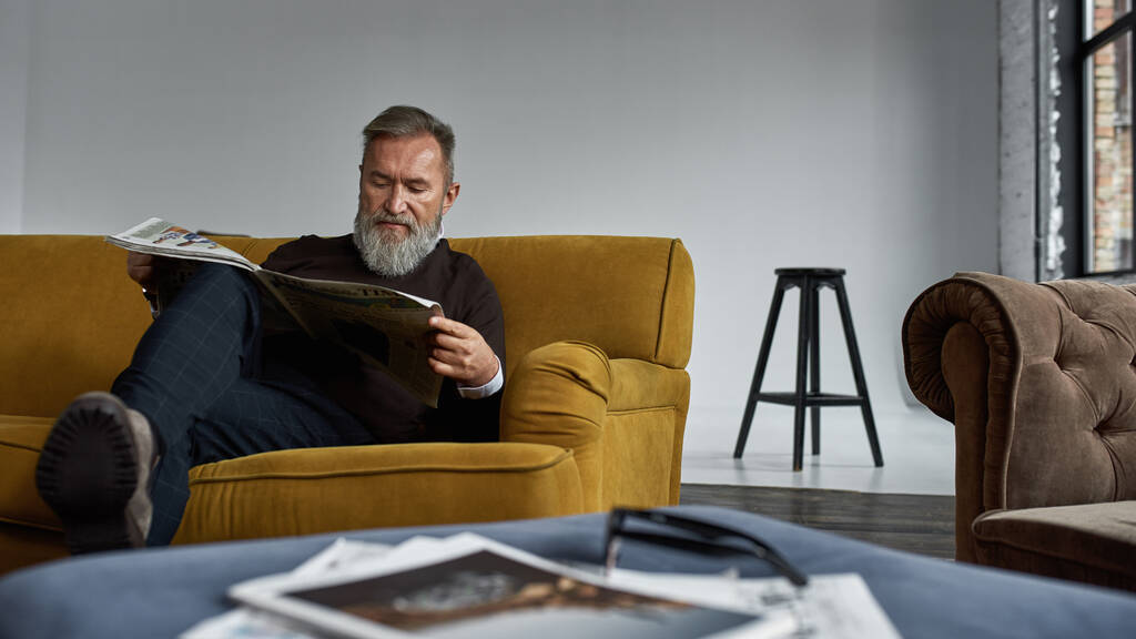 Elderly concentrated caucasian man reading newspaper on sofa in spacious apartment. Trendy bearded pensioner wearing casual clothes. Modern senior male lifestyle. Domestic rest and leisure - Fotografie, Obrázek
