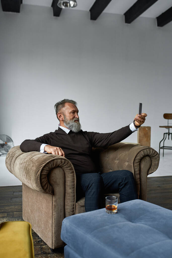 Kafkasyalı yaşlı bir adam dinleniyor ve geniş bir dairedeki koltukta akıllı telefondan selfie çekiyor. Moda sakallı emekli. Modern son sınıf erkek yaşam tarzı. Ev eğlencesi ve eğlence. - Fotoğraf, Görsel