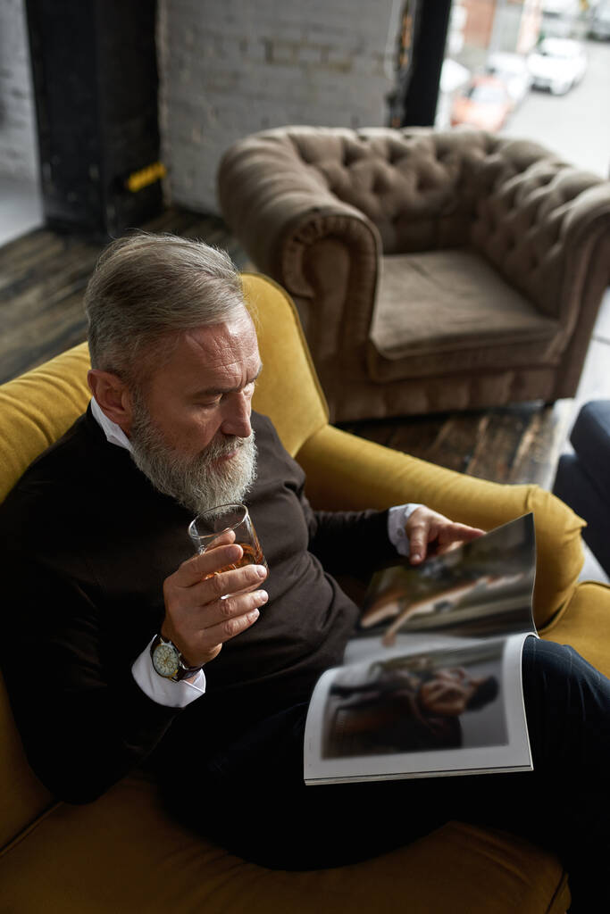 Top view of senior focused caucasian man drinking brandy and reading journal on sofa in spacious apartment. Stylish bearded pensioner. Modern elderly male lifestyle. Domestic rest and leisure - Foto, Bild