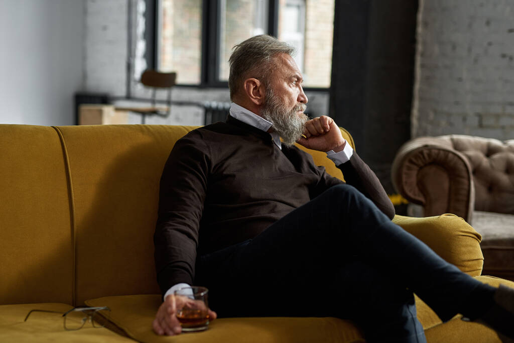 Elderly thoughtful caucasian man drinking cognac and looking away on sofa in spacious apartment. Trendy bearded pensioner. Modern senior male lifestyle. Domestic rest and free time - Zdjęcie, obraz