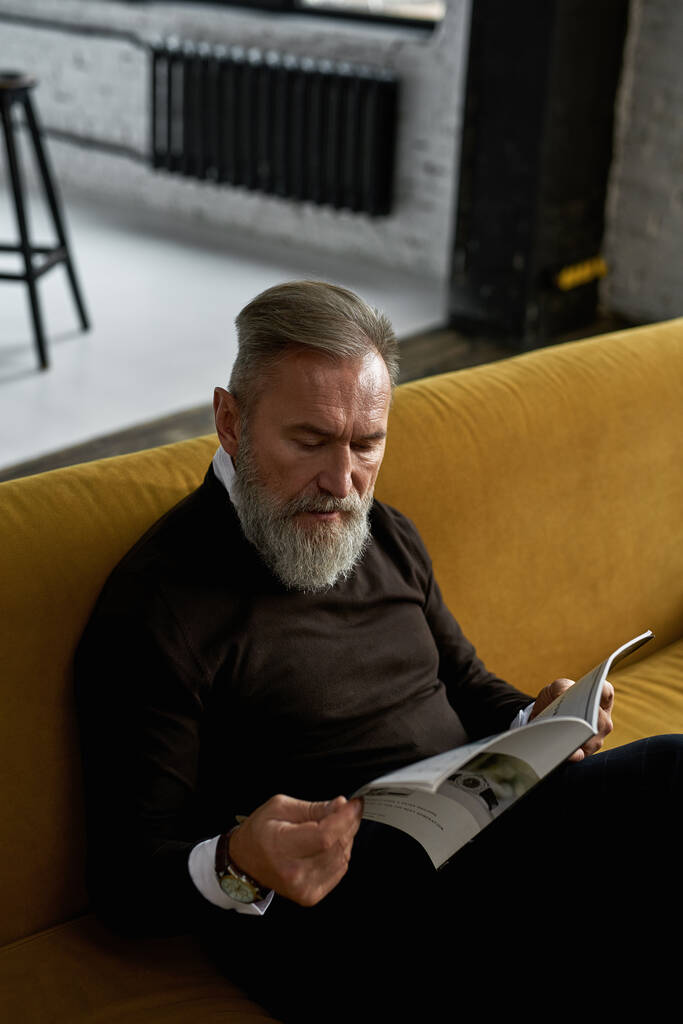 Elderly concentrated caucasian man reading journal on sofa in spacious apartment. Trendy bearded pensioner wearing casual clothes. Modern senior male lifestyle. Domestic rest and leisure - Foto, Imagem