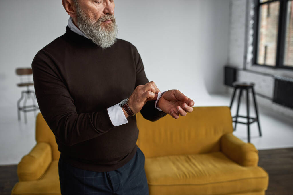 Obscure face of elderly serious caucasian man correcting shirt sleeve in spacious apartment. Trendy bearded pensioner wearing casual clothes. Concept of modern senior male lifestyle. Daytime - Photo, Image