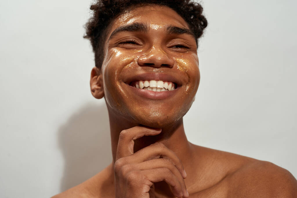 Getroffen van een lachende zwarte man met een gouden masker op zijn gezicht. Jonge brunette krullende man met naakte romp. Concept van gezichtshuid verzorging. Geïsoleerd op witte achtergrond. Studiosessie - Foto, afbeelding