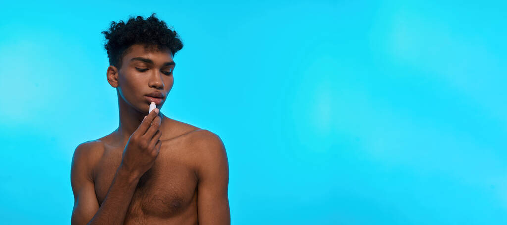 Partial image of black guy painting lips with hygienic lipstick. Young brunette curly man with naked torso. Body and skin care. Isolated on light blue background. Studio shoot. Copy space - Photo, image