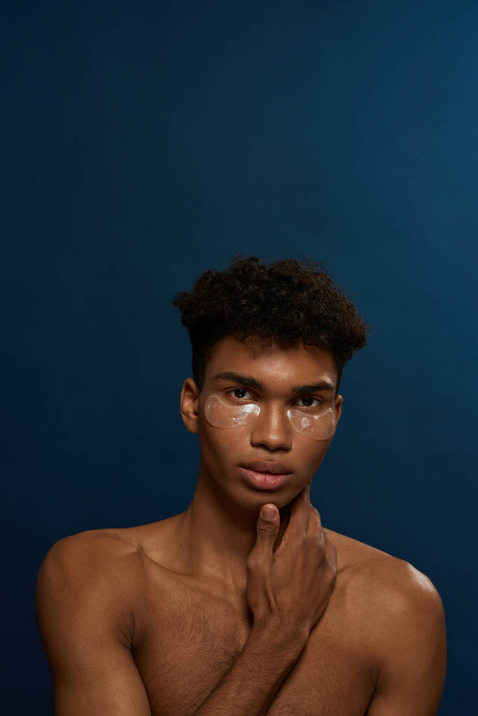 Portrait of confident black guy with under eye patches on face looking at camera. Young man with naked torso. Body and skin care. Facial lifting and rejuvenation. Blue background in studio. Copy space - Zdjęcie, obraz