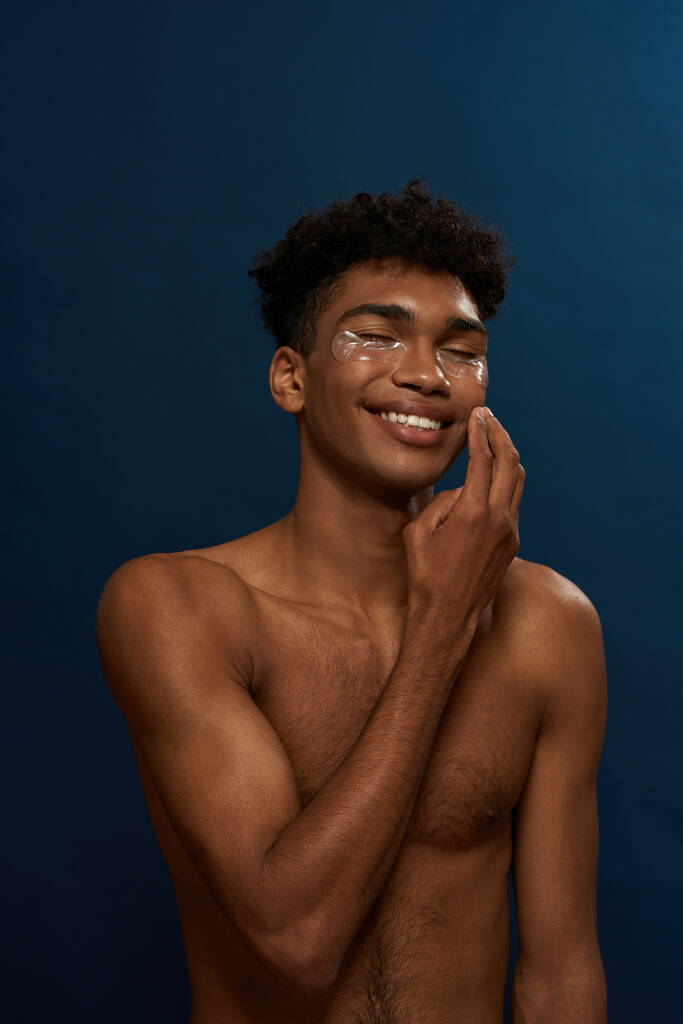Cropped of smiling black guy correct under eye patch on face. Young man with naked torso and closed eyes. Body and skin care. Facial lifting and rejuvenation. Blue background. Studio shoot. Copy space - Foto, imagen