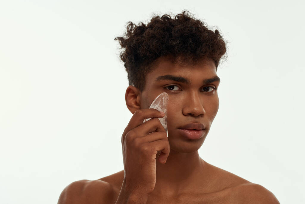 Cropped image of black guy applying ice slice on his face. Young brunette curly man with naked torso looking at camera. Body and skin care. Isolated on white background. Studio shoot. Copy space - Φωτογραφία, εικόνα