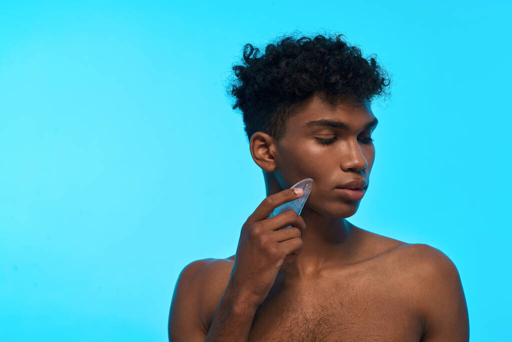 Partial image of black guy applying ice slice on his face. Young slim brunette curly man with naked torso. Body and skin care. Isolated on light blue background. Studio shoot. Copy space - Fotó, kép