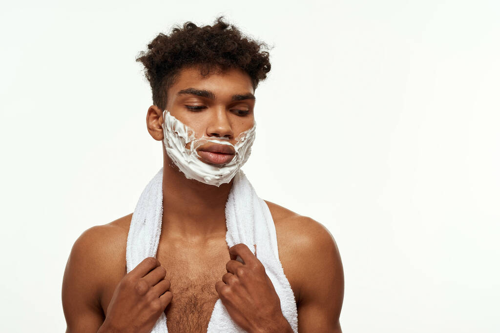 Cropped of thoughtful black guy with shaving foam on his face. Young brunette curly man with towel and naked torso. Hygiene and skin care. Isolated on white background. Studio shoot. Copy space - Valokuva, kuva