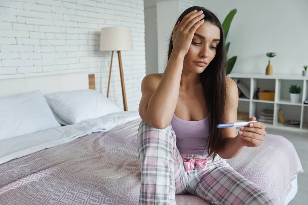Frustrated young woman holding pregnancy test and touching head while sitting on bed at home - Foto, immagini