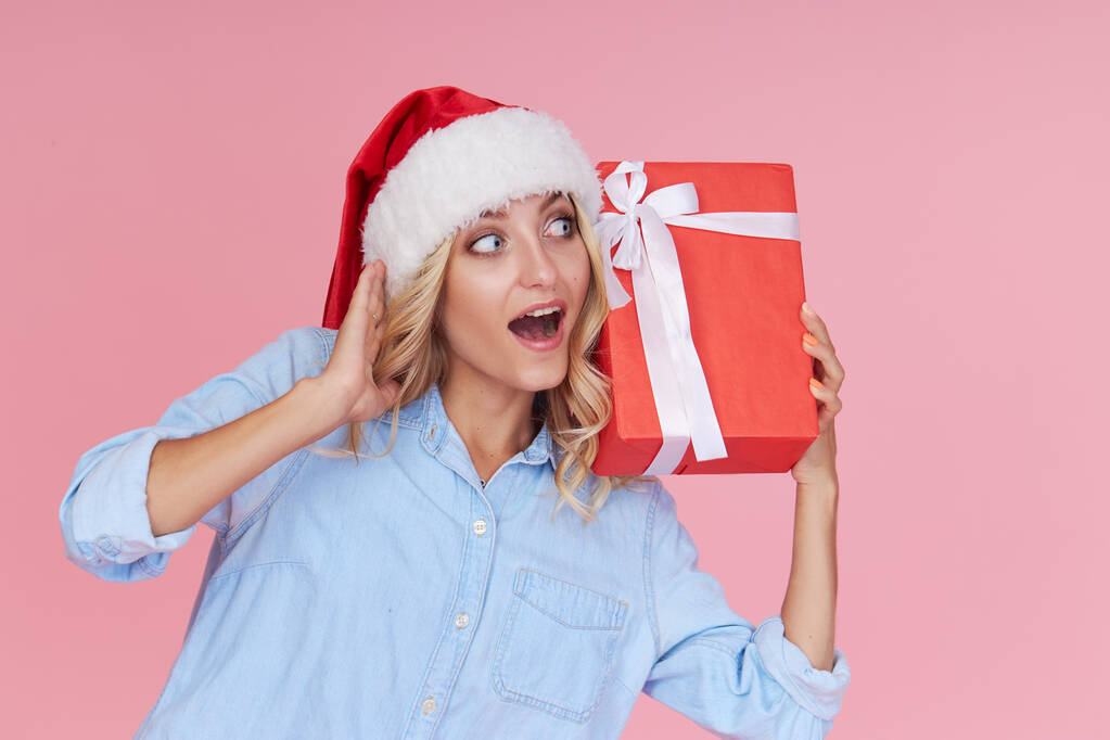Portrait of a cheerful joyful girl in casual clothes, Santa hat with a red gift box on a pink background. Wow face. Concept of Christmas and New year holidays - Фото, изображение
