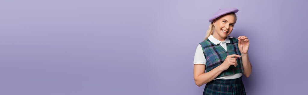 Happy blonde student in beret holding eyeglasses on purple background, banner   - Foto, Imagen