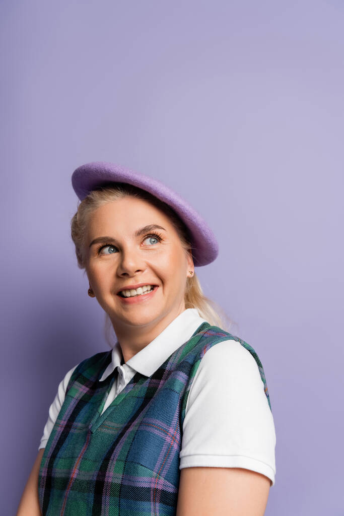 Smiling student in checkered vest and beret looking away on purple background  - Foto, immagini
