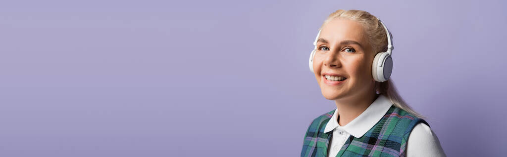 Positive blonde student in headphones looking at camera isolated on purple, banner  - Foto, immagini