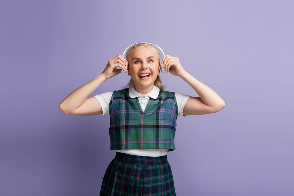 Positive student in uniform holding headphones near ears isolated on purple  - Foto, Imagem