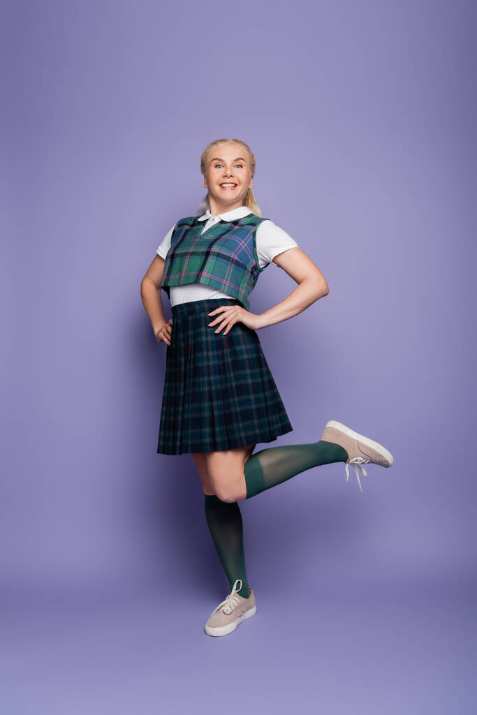Positive student in checkered uniform holding hands on hips on purple background  - Фото, изображение
