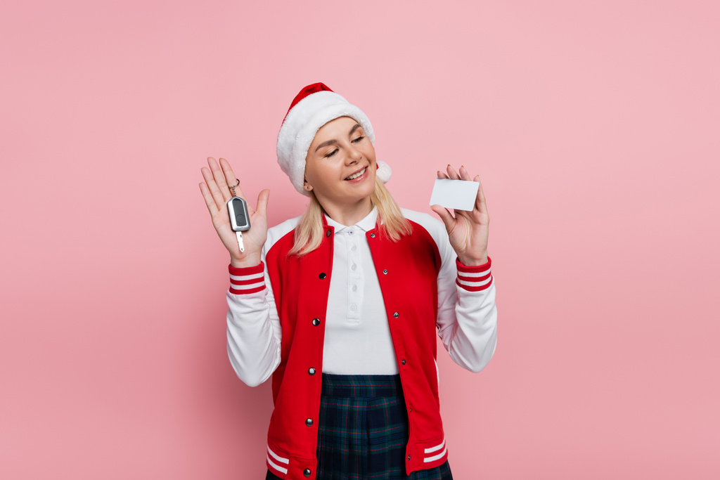 Cheerful woman in santa hat holding car key and empty driving license isolated on pink  - Photo, Image