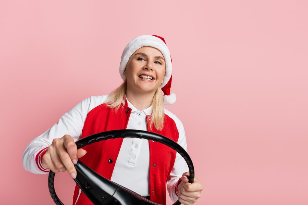 Smiling woman in santa hat holding steering wheel isolated on pink  - Fotoğraf, Görsel