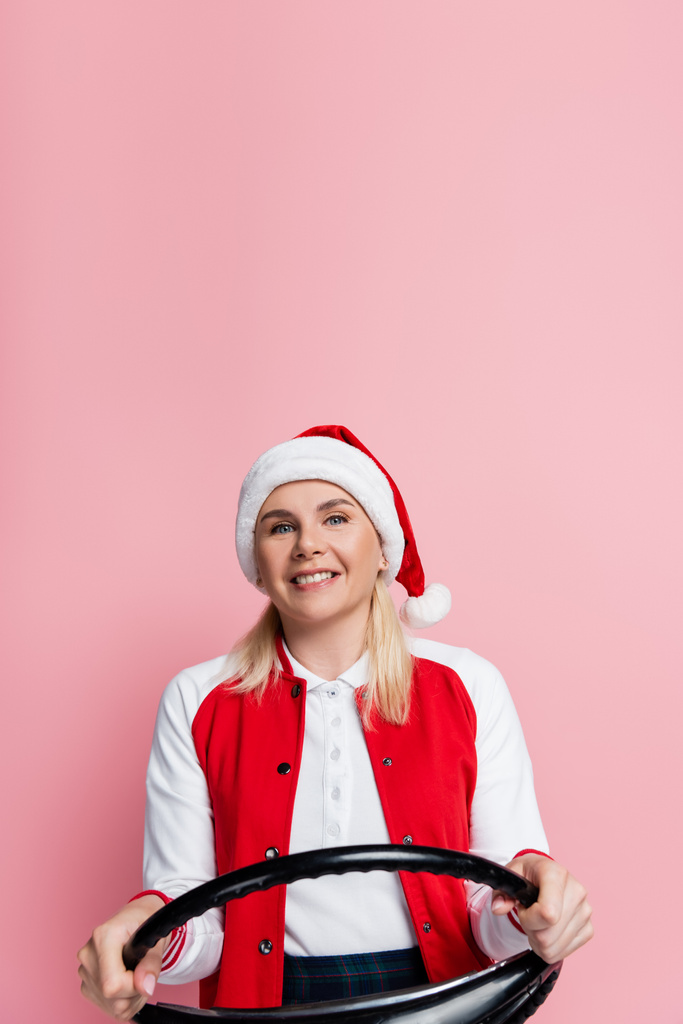 Cheerful woman in red santa hat holding steering wheel isolated on pink  - Foto, immagini