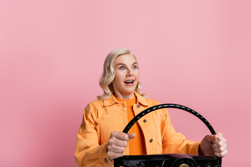 Shocked blonde driver holding steering wheel isolated on pink  - Foto, Bild