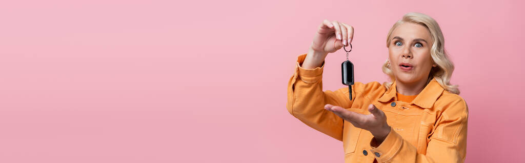 Amazed blonde woman pointing at car key isolated on pink, banner  - Foto, afbeelding