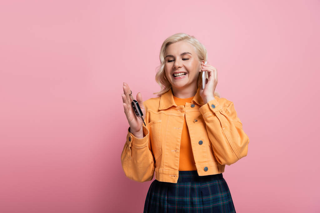 Happy blonde woman holding car key and talking on mobile phone isolated on pink  - Foto, Imagen