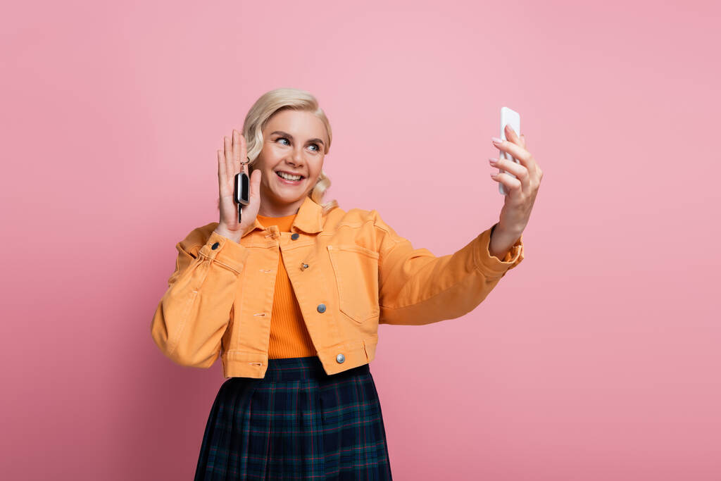 Smiling woman taking selfie on smartphone while holding car key isolated on pink  - Foto, immagini