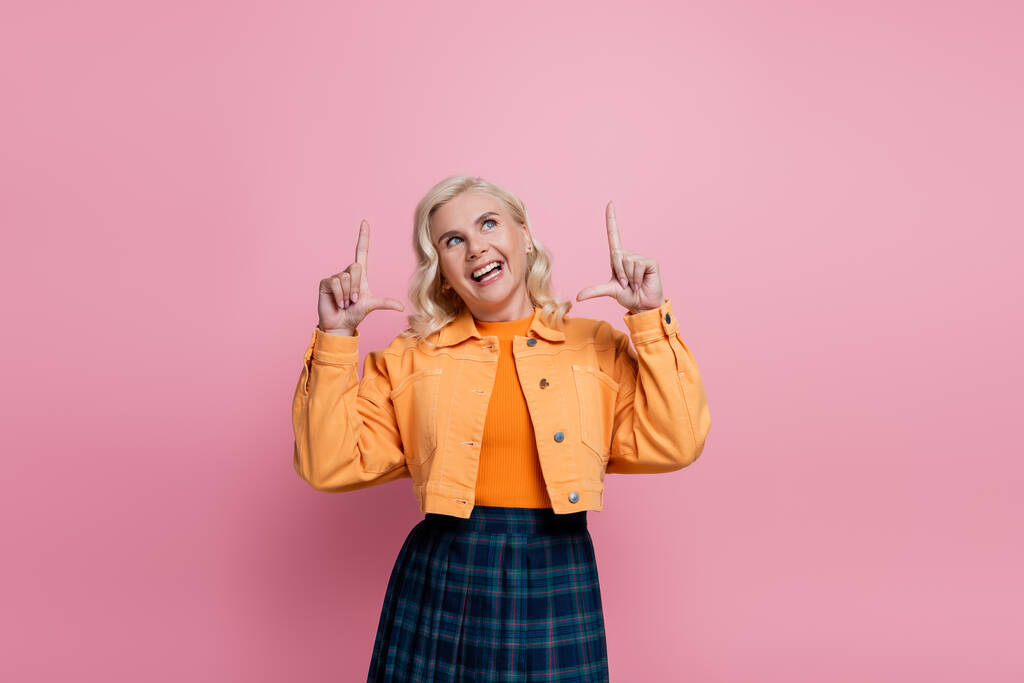 Positive woman in orange jacket pointing with fingers and looking up isolated on pink  - Foto, Bild