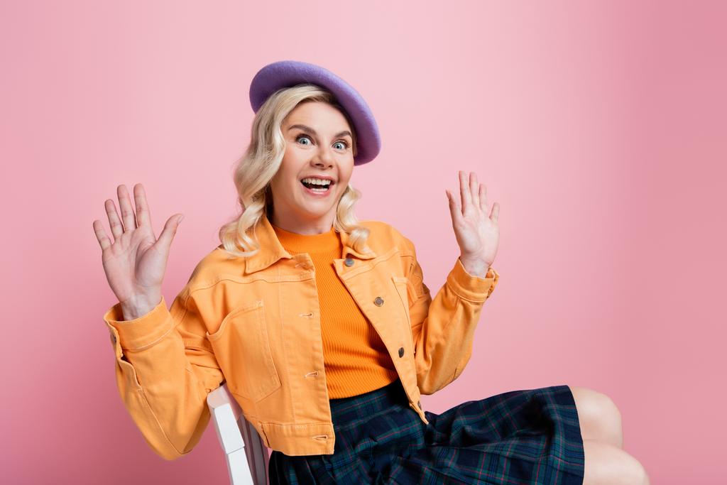 Astonished blonde woman in beret sitting on chair isolated on pink  - Foto, immagini