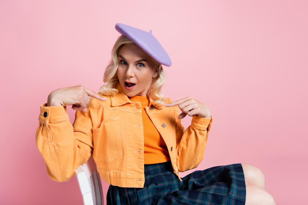 Shocked woman in beret pointing with fingers while sitting on chair isolated on pink  - Photo, image