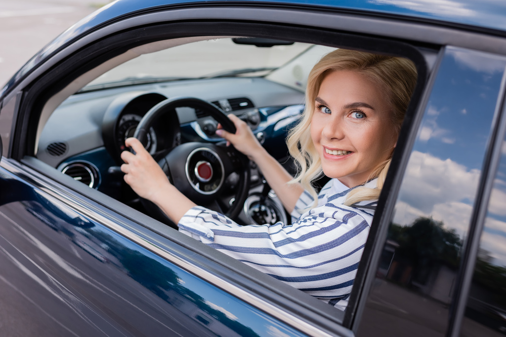 Blonde woman looking at camera in blurred car during driving course  - Foto, Bild