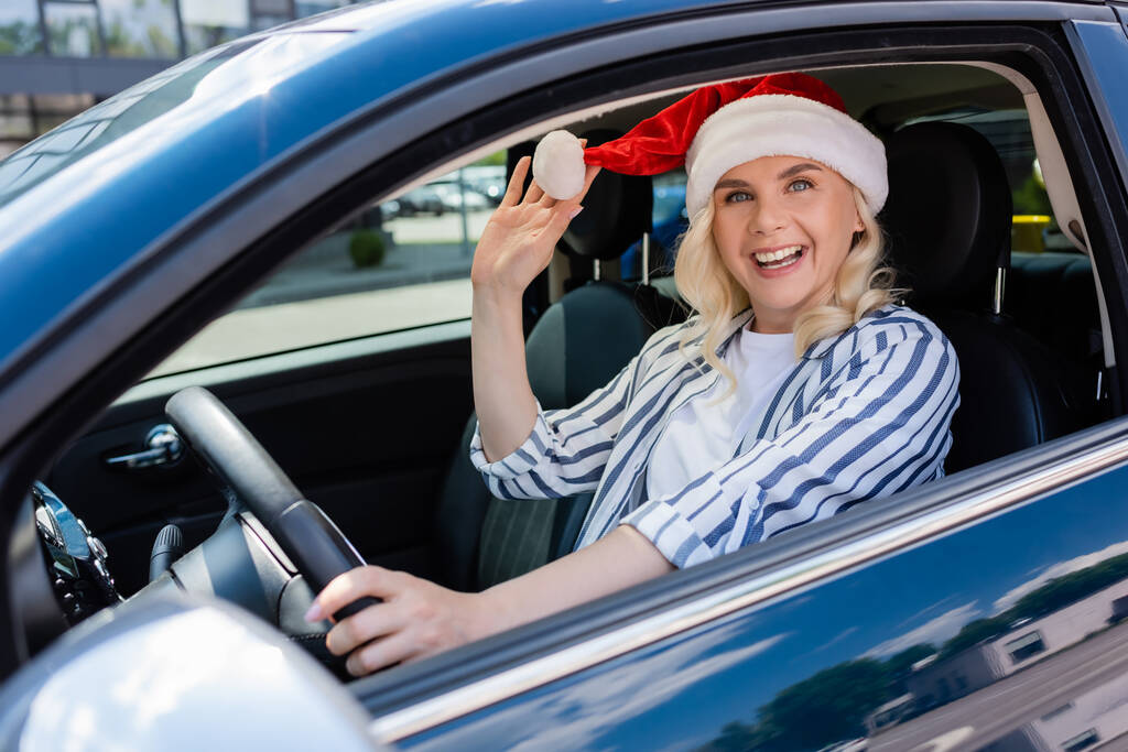 Positive blonde driver in santa hat looking at camera while driving car  - Photo, Image