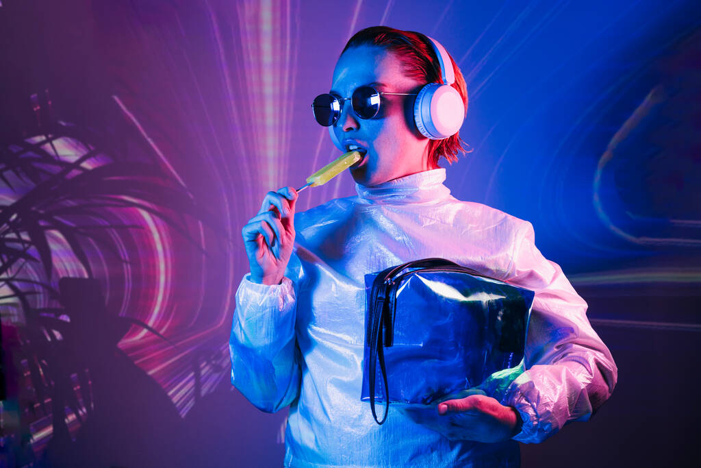 Portrait of a cyberpunk girl in sunglasses and headphones in neon light. Futuristic woman licking a lollipop in holographic clothes on the background of waves Concept. fashion, futuristic generation - Fotografie, Obrázek
