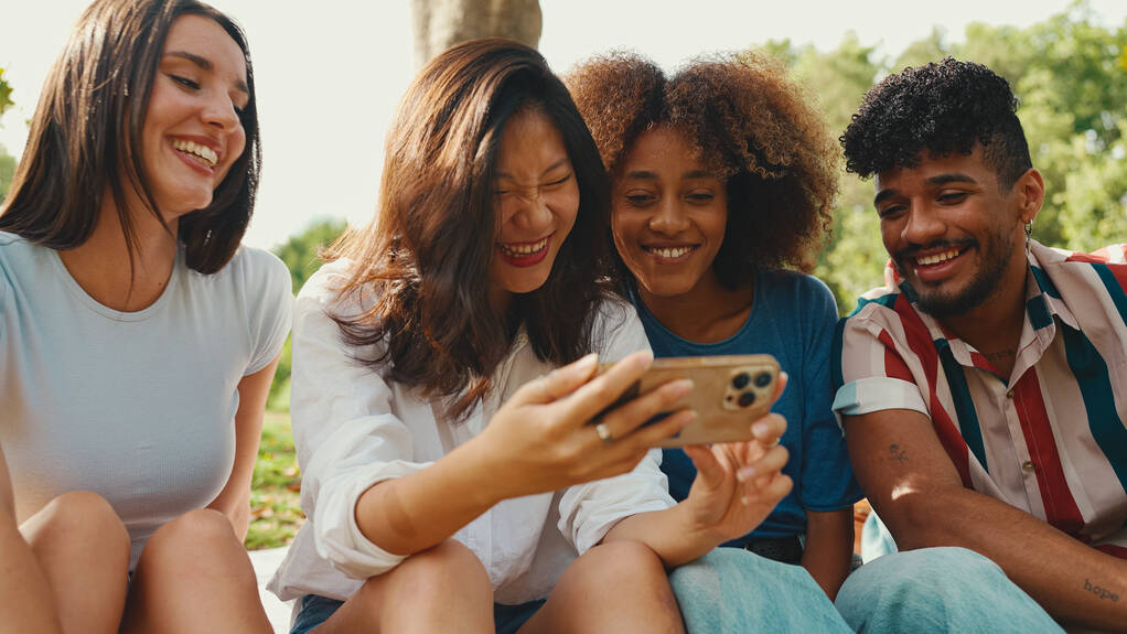 Happy, smiling multiethnic young people at picnic on summer day outdoors. Group of friends talking, using cellphone while relaxing in the park at picnic - 写真・画像