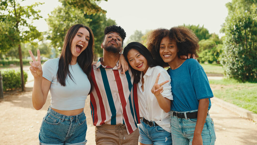Щасливі, милі багатоетнічні молоді люди позують на камеру в літній день на відкритому повітрі. група друзів обіймає один одного посміхаючись на камеру, стоячи на шляху в парку
 - Фото, зображення
