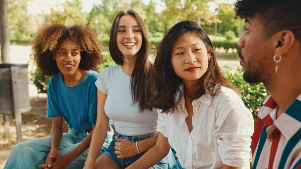 Jóvenes multiétnicos felices hablando mientras están sentados en el banco del parque en el día de verano al aire libre. Grupo de amigos hablando y riendo alegremente en el parque de la ciudad - Foto, imagen