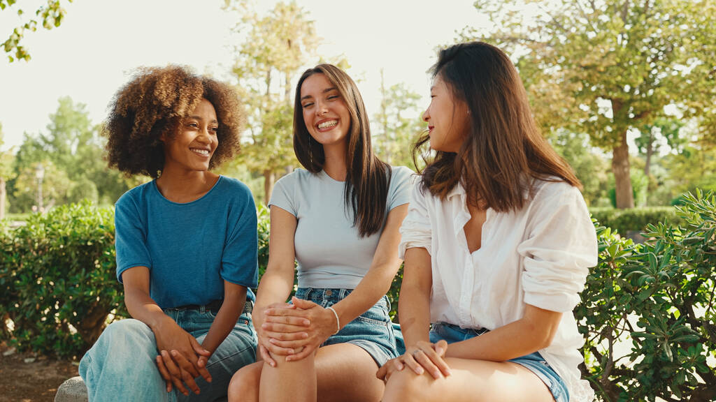 Щасливі багатоетнічні молоді жінки розмовляють, сидячи на лавці в літній день на відкритому повітрі. Група дівчат, що розмовляють і весело сміються в міському парку
 - Фото, зображення