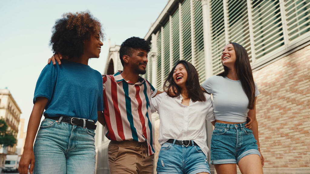 Šťastní multietničtí mladí lidé chodí objímat v letní den venku. Skupina přátel mluví a vesele se směje, když jde po ulici. - Fotografie, Obrázek