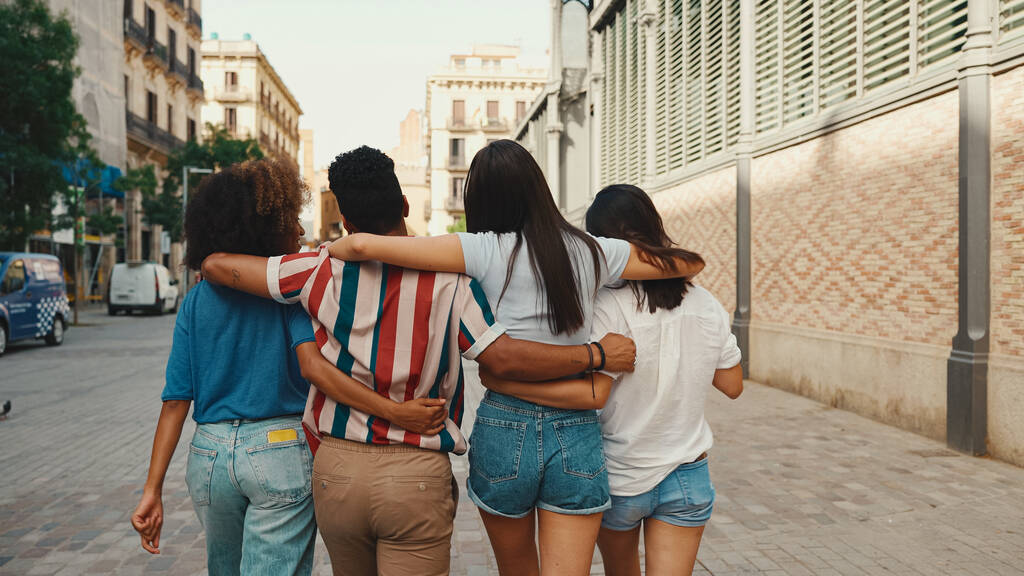 Boldog többnemzetiségű fiatalok sétálnak ölelkezve a nyári napon a szabadban. A baráti társaság vidáman beszél és nevet, miközben sétálnak az utcán. - Fotó, kép