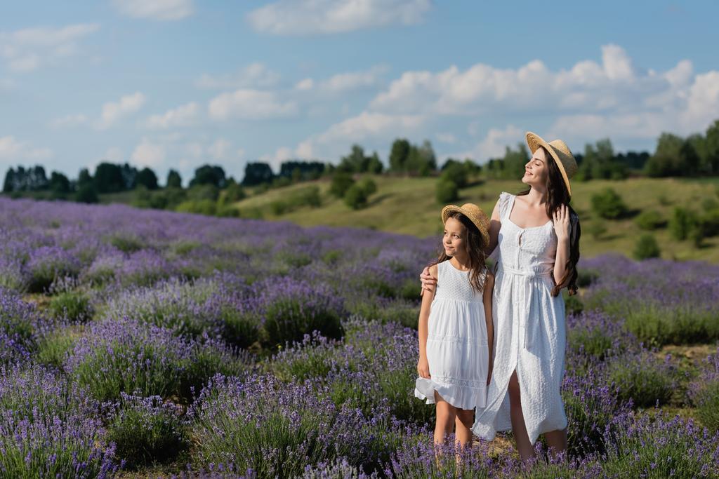smiling woman in straw hat and white dress embracing daughter in lavender meadow - Photo, Image