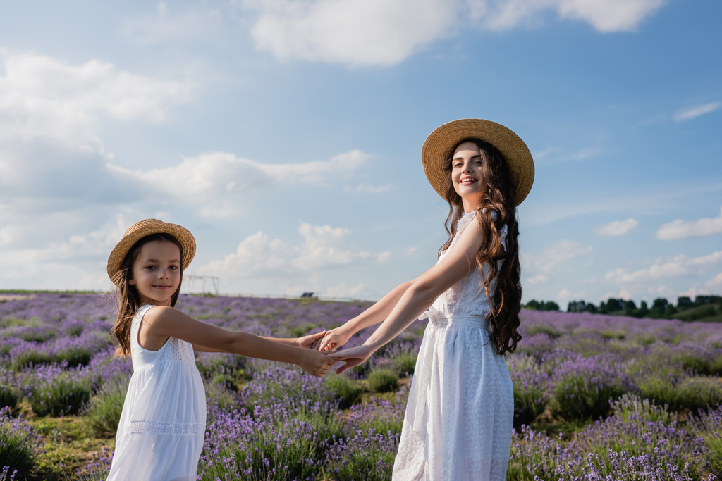 cheerful woman and girl in straw hats holding hands and looking at camera in meadow - Photo, Image