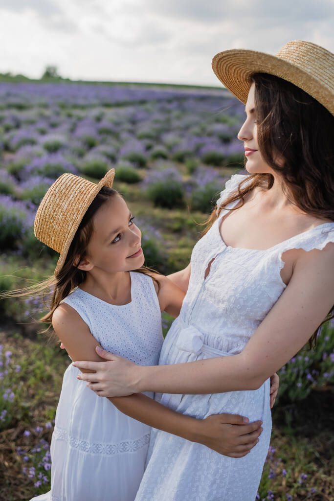 mom and daughter in straw hats smiling at each other in flowering field - Foto, Imagen
