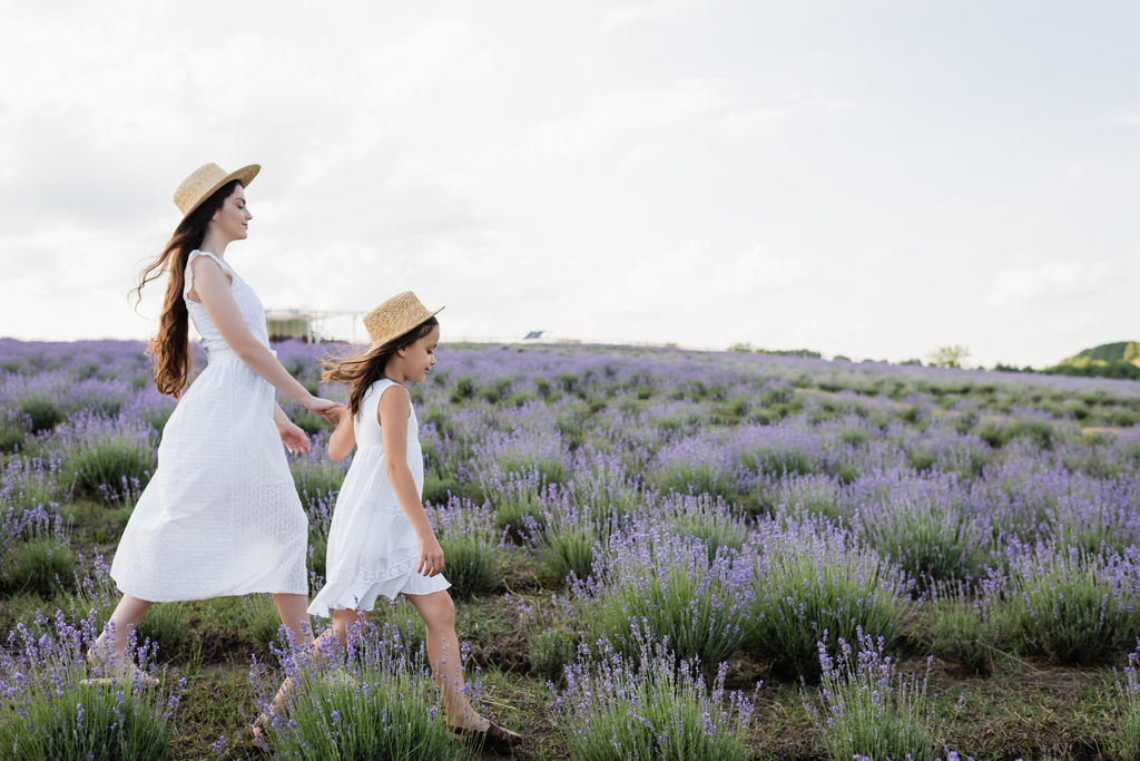 side view of mom and child in white dresses and straw hats holding hands and walking in field - Foto, Imagem