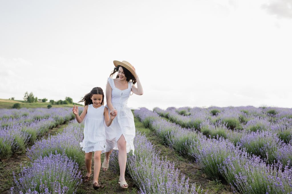cheerful mother and daughter in white dresses holding hands while running in meadow - Fotografie, Obrázek