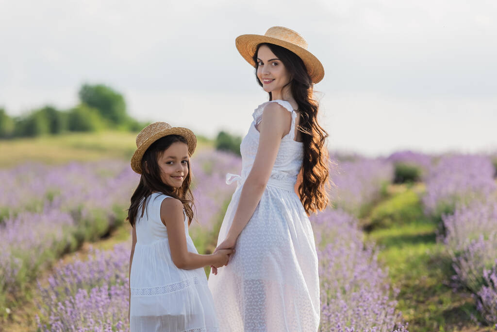 joyful girl and woman with long hair looking at camera and holding hands in meadow - 写真・画像