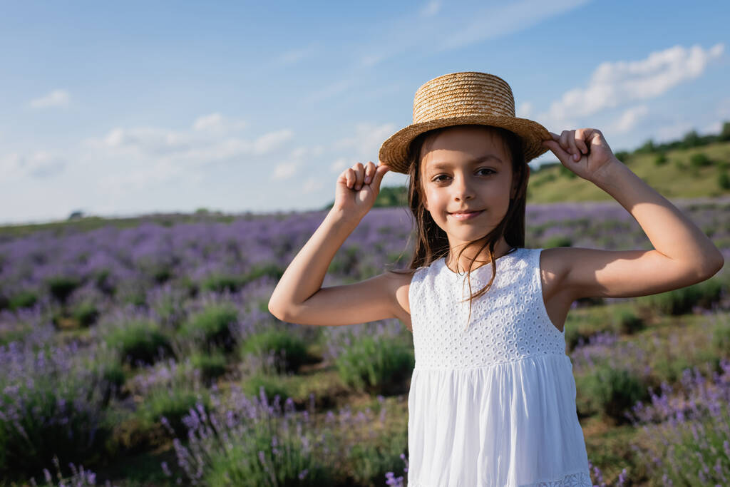 girl smiling at camera while adjusting straw hat in blurred lavender field - Foto, afbeelding