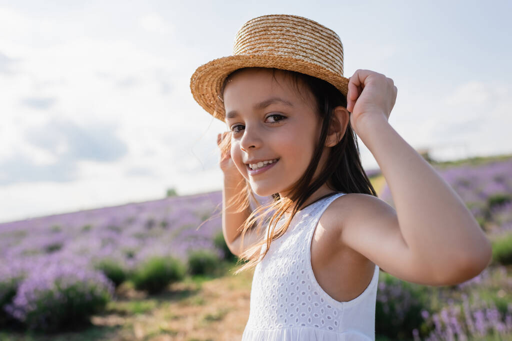 pleased girl in straw hat looking at camera in blurred lavender meadow - Foto, Imagem
