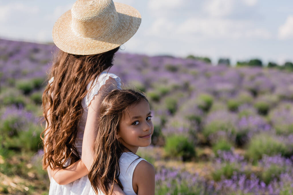 счастливый ребенок смотрит на камеру рядом с мамой в соломенной шляпе в размытом поле - Фото, изображение