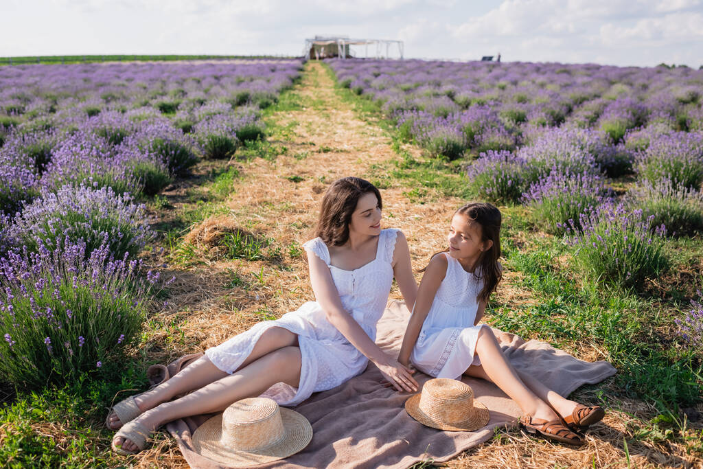 mother and daughter in white dresses looking at each other near straw hats on blanket in meadow - Fotografie, Obrázek